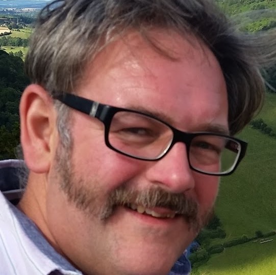 Smiling head-and-shoulders portrait of Alan Coxon wearing black glasses, with a green countryside landscape in the background.