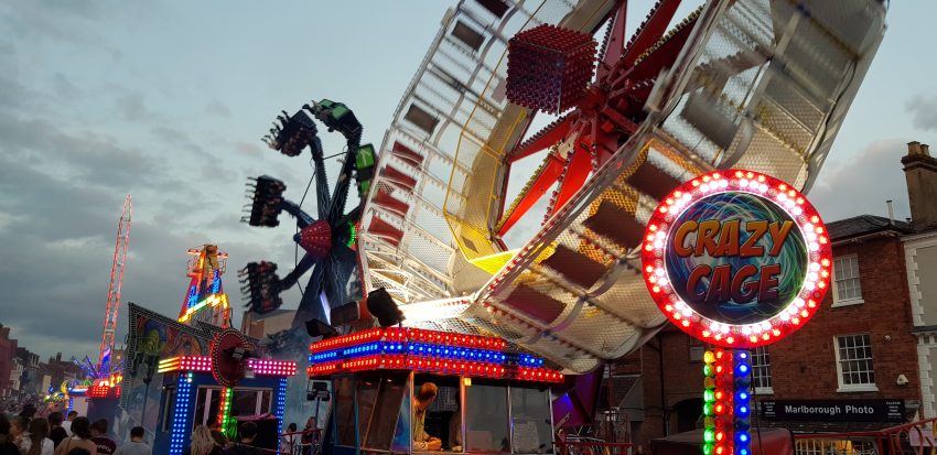 A lively evening scene at Marlborough Mop Fair, with brightly lit funfair rides against a cloudy twilight sky. In the foreground, the illuminated sign for the "Crazy Cage" ride glows with multicoloured lights, while the ride itself spins dramatically. Other fairground rides, colourful lights, and crowds of people enjoying the event fill the background along the High Street, with traditional brick buildings lining the scene.