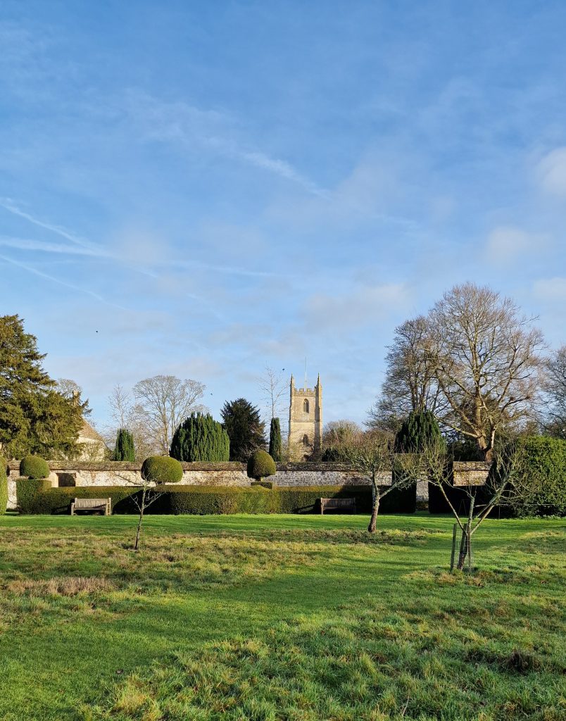 Winter sunlight over a manicured garden with clipped hedges, benches and a stone church tower beyond a low wall.