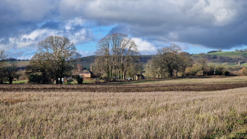 Winter farmland in Wiltshire with bare trees, stubble fields and dramatic clouds over rolling hills.