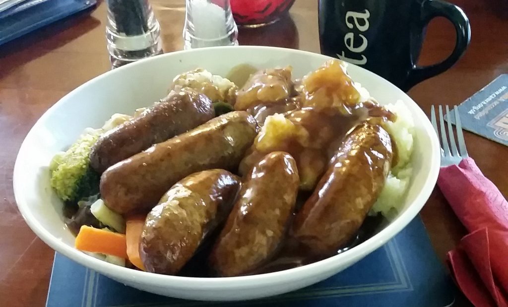 Bowl of sausages with gravy served on mashed potatoes and seasonal vegetables, with a mug of tea on a pub table.