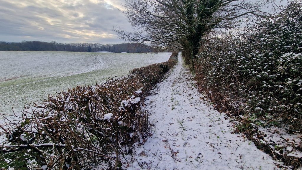 Snow-covered footpath running beside hedges and open fields in the winter countryside.