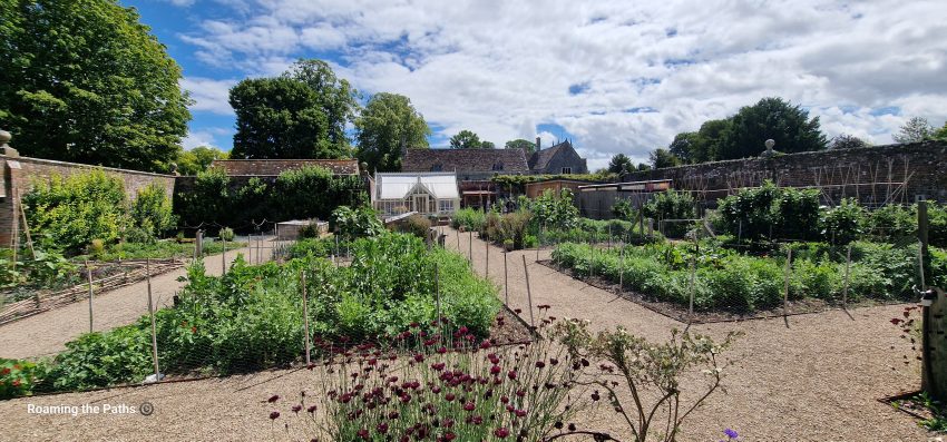 Productive kitchen garden at Avebury Manor with vegetable beds, gravel paths, a glasshouse, and historic stone walls beneath a blue summer sky.